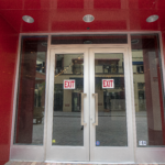 Double glass exit doors with 'EXIT' signs, framed by a red facade, showcasing Oratso Corporation's door installations at Sunset Place Mall.