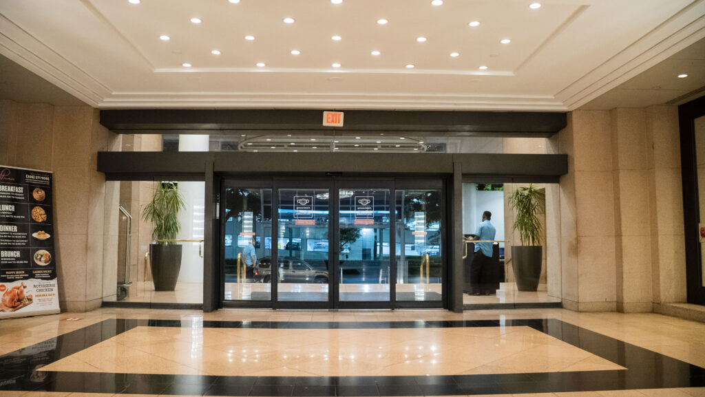 Automatic sliding glass doors at Oratso Corporation's apartment complex entrance, featuring a modern interior with polished marble flooring and decorative plants.
